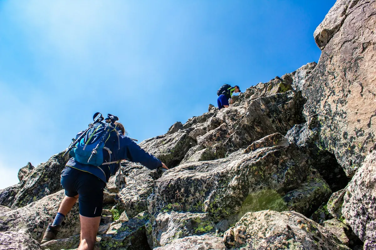 Two hikers with backpacks climbing a rocky mountain trail under a clear blue sky.