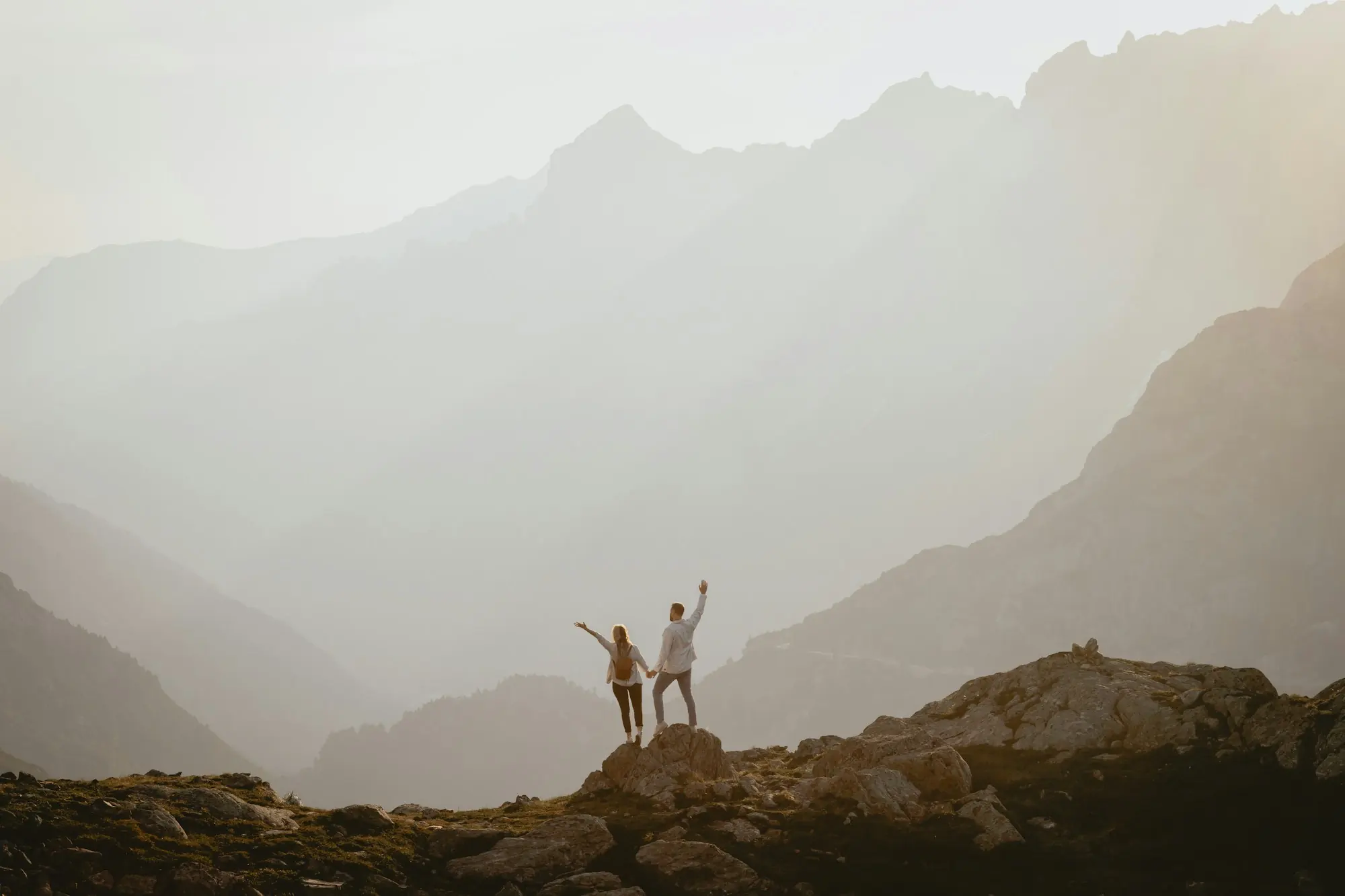 Two small figures standing on a rocky summit in soft golden light, with layers of misty mountain ridges behind them.