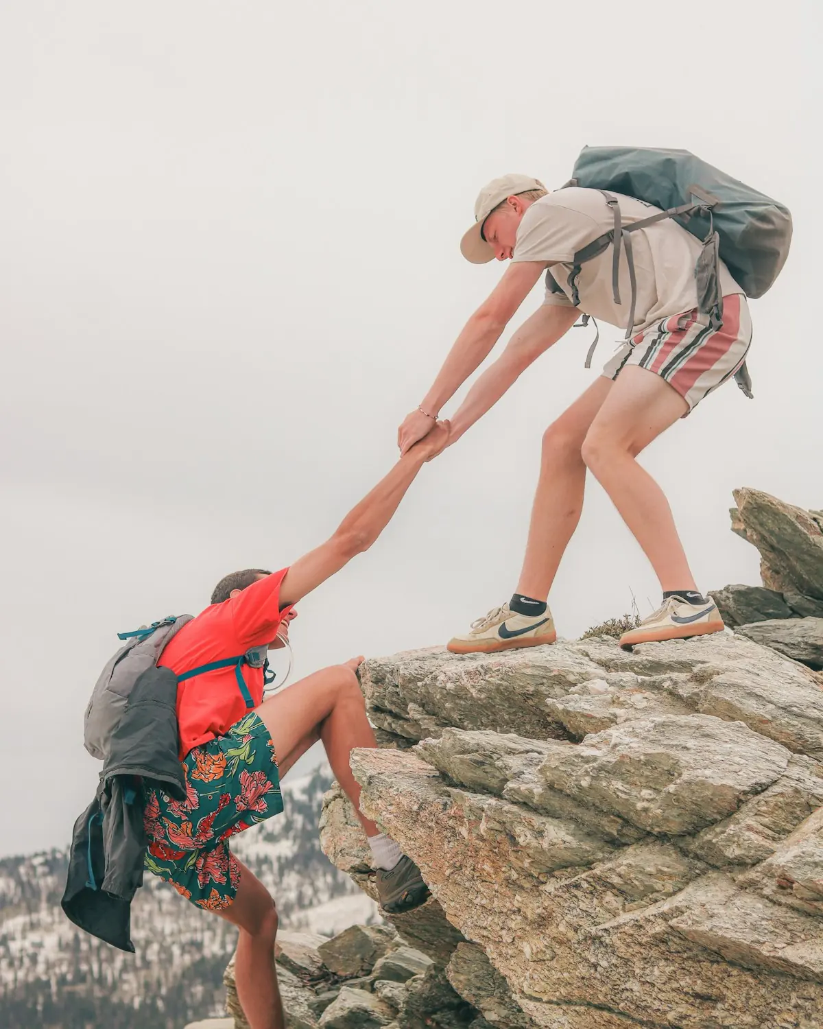 A hiker standing on a rocky outcrop grips the hand of another hiker and pulls him up the last step, with snowy forested hills below.